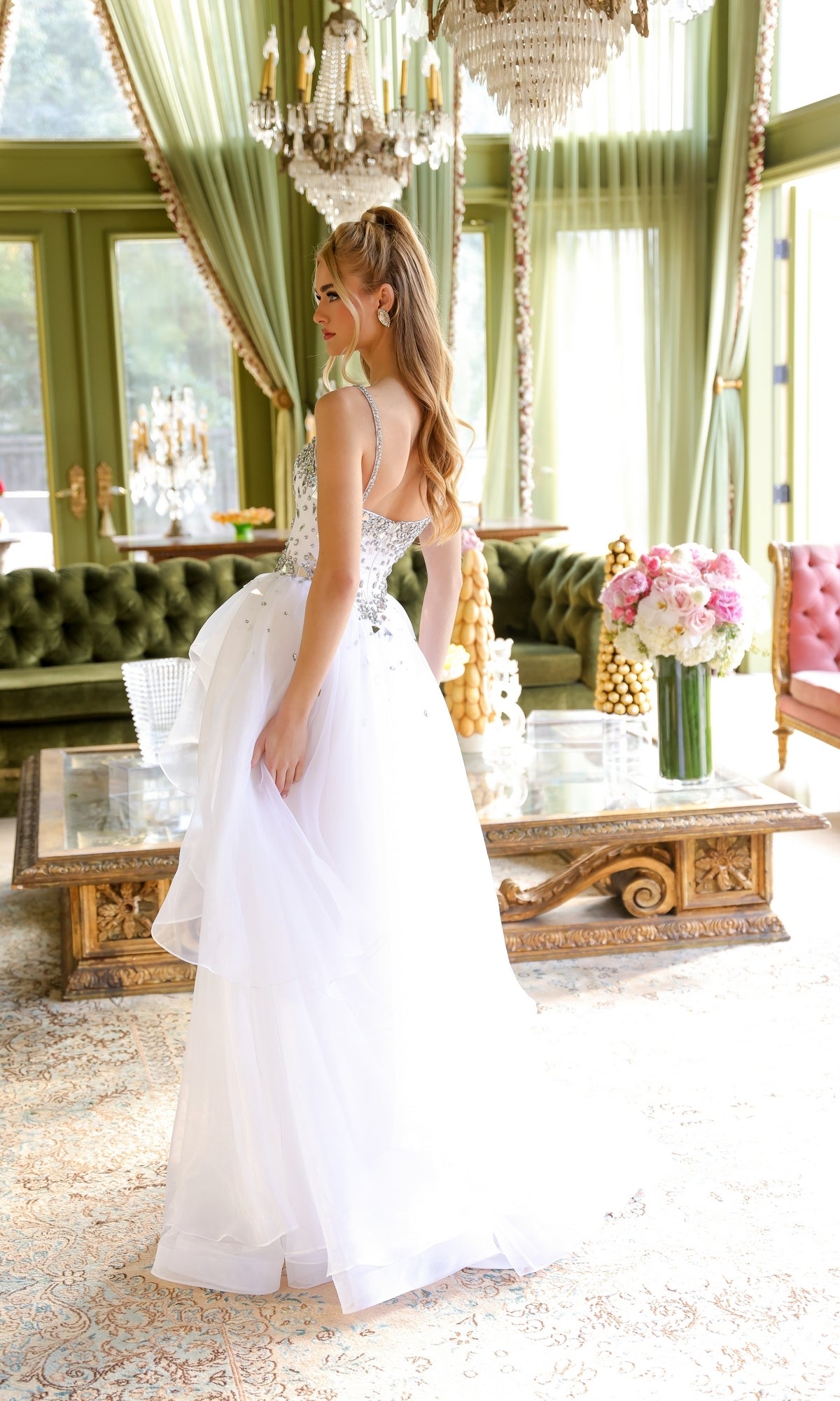 A woman stands in an elegant room with green curtains, a chandelier, and ornate furniture, wearing the Ava Presley 29189 Tiered Long Prom Ball Gown and looking over her shoulder.